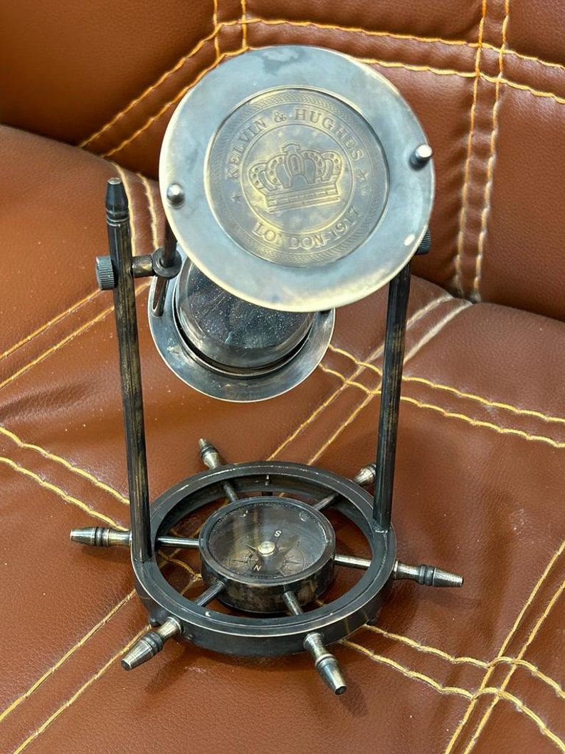 Op de afbeelding: Een zandloper van messing met een kompasvoet. De zandloper heeft een kroon en de tekst "Kelvin & Hughes London 1917" op de bovenkant. De kompasvoet heeft een scheepsrad-ontwerp.
