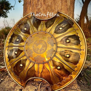 Pode incluir: Um escudo grande, redondo e dourado com um design decorativo. O escudo tem um design circular central com um padrão floral e é rodeado por oito seções radiantes com uma borda festoneada. O escudo está apoiado no chão em frente a um tronco de árvore.