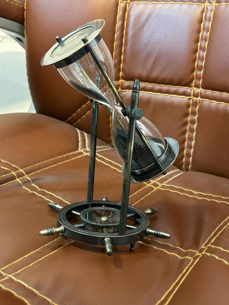 Op de afbeelding: Een zwarte metalen zandloper met een kompasvoet. De zandloper is gevuld met zwart zand en heeft een heldere glazen bol. De kompasvoet heeft een zwart metalen wiel met spaken.