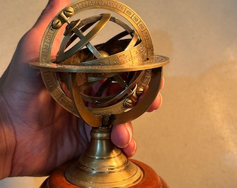 May include: A brass celestial globe on a wooden base. The globe is marked with degrees and has a compass rose. The globe is on a wooden base with the text "TheCraftEra" on it.
