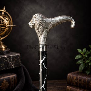 May include: A walking stick featuring a silver-coloured handle shaped like a lion's head with detailed mane textures. The shaft is black with white marble-like veining. The text TheCraftEraShop is visible on the handle. The design is metallic.