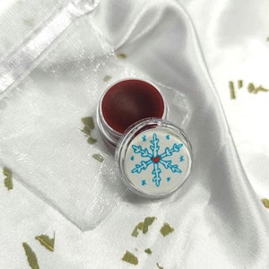 Puede incluir: Un pequeño recipiente redondo de bálsamo labial rojo con una tapa blanca que presenta un copo de nieve azul con un corazón rojo en el centro. El recipiente está sobre una superficie blanca con formas de hojas verdes.