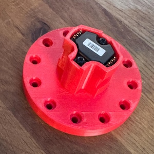 Może przedstawiać: Jasnoczerwony, drukowany w 3D komponent z okrągłą podstawą i centralną, wieloaspektową strukturą. Podstawa posiada równomiernie rozmieszczone otwory. Czarny prostokątny moduł z naklejką z kodem kreskowym jest zamocowany pośrodku.