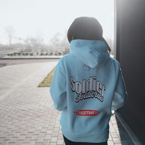 Könnte beinhalten: Hellblauer Kapuzenpullover mit dem Wort "Soldier" in einer stilisierten Schriftart auf dem Rücken, zusammen mit dem Wort "Streetwear" in einem roten Oval. Die Person trägt einen schwarzen Hut.