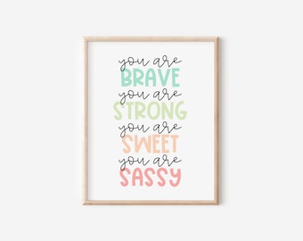 以下が含まれることがあります： 「you are BRAVE, you are STRONG, you are SWEET, you are SASSY」という言葉がカラフルで遊び心のあるフォントで書かれた額入りのプリント。ミントグリーン、ライトグリーン、ピーチ、ピンクのパステルカラーを使用。フレームは明るい木の色。