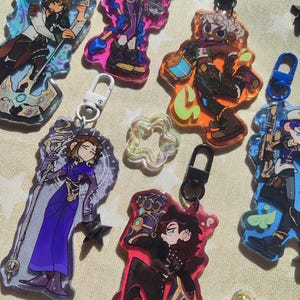 Può includere: Sei ciondoli colorati con personaggi con accenti glitter e portachiavi attaccati. I ciondoli presentano vari personaggi con diversi stili di capelli, vestiti e accessori. I personaggi sono raffigurati in uno stile cartone animato.