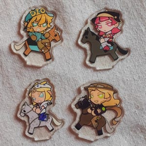 May include: Four clear acrylic charms depicting cartoon characters on horses. Each charm showcases a distinct character design with varied outfits and hair colours. The charms are set against a light-coloured fabric backdrop.