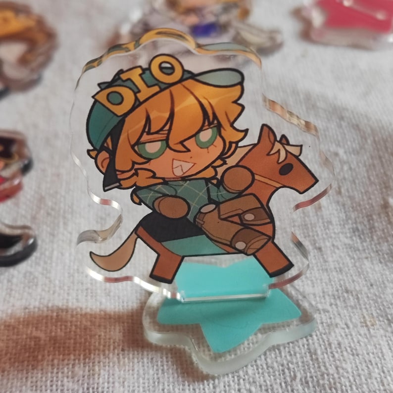 May include: A clear acrylic charm depicting a cartoon character with orange hair and a green cap, riding a brown stick horse. The charm has the word "DIO" on the cap and is mounted on a light blue base.