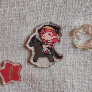 May include: A clear acrylic charm depicting a cartoon character with pink hair and a black hat riding a grey horse. A red star-shaped charm and a cloud-shaped charm are also visible.