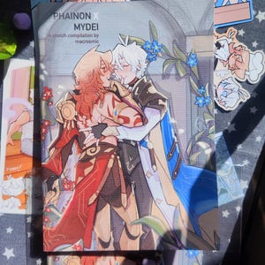以下が含まれることがあります： 「PHAINON X MYDEI」というタイトルのスケッチコンピレーションブック。2人のキャラクターが抱き合っているアニメスタイルのイラストが特徴です。表紙は赤、金、青のアクセントが施されています。本の周りには、いくつかのキャラクターのステッカーが見えます。