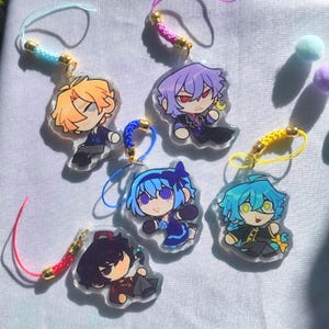 Può includere: Una collezione di ciondoli in acrilico con personaggi in stile anime con capelli e abiti colorati. Ogni ciondolo presenta un design di personaggio diverso ed è attaccato a un cordino colorato con una chiusura dorata. I ciondoli sono su una superficie bianca.