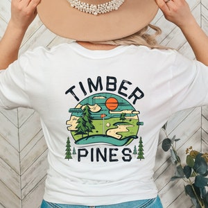 May include: White t-shirt with a green, blue, and orange graphic of a golf course with the text "Timber Pines" in a circle.