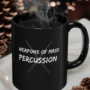 May include: A black ceramic mug with the text "WEAPONS OF MASS PERCUSSION" in white, along with a graphic of crossed drumsticks. Steam rises from the dark liquid inside. The mug has a curved handle and is set against a blurred background.