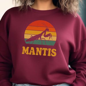 May include: Burgundy sweatshirt featuring a retro design of a praying mantis silhouette set against a sunset gradient of orange, yellow, and green. The word "MANTIS" is in a bold, vintage font below the insect.