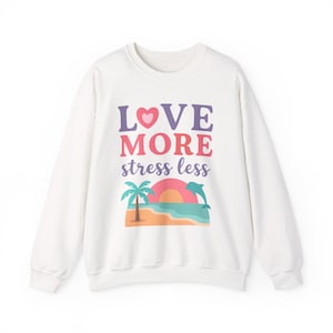 May include: White sweatshirt with the words "LOVE MORE stress less" in purple, pink, and blue. A graphic of a palm tree, sunset, and dolphin is below the text. The sweatshirt is a crew neck style.
