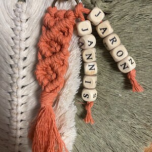 Puede incluir: Un llavero de macramé de color coral con un borla. El llavero tiene dos juegos de cuentas de madera con los nombres "Jaron" y "Jannis" escritos en negro.