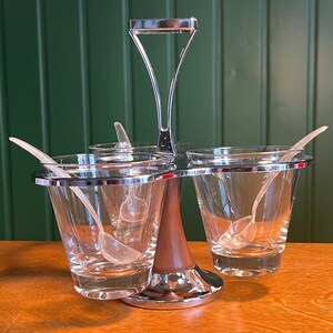 May include: A clear glass condiment server with a silver-colored metal rim and handle. The server has three glass cups, each with a small plastic spoon. The base is a combination of wood and metal.