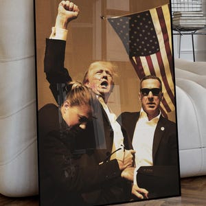 May include: A black and white photo of three people. The person in the center is raising their fist in the air. The person on the left is wearing a black jacket and the person on the right is wearing a black suit. The background is a blurry image of an American flag.