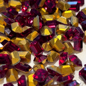 May include: A collection of faceted, geometric-shaped gemstones. The gemstones are a deep ruby red and gold, with a metallic finish. The shapes vary, including rectangular and hexagonal forms. The gemstones are scattered across a light-colored surface.