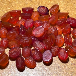 May include: A pile of 40 oval-shaped carnelian gemstones with a carved design. The stones are a deep orange-red colour and have a translucent finish.