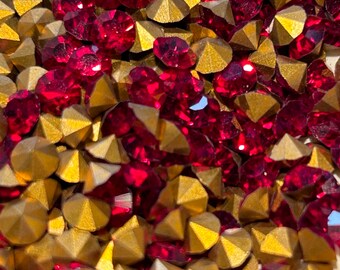 May include: A close-up image of a pile of red and gold rhinestones. The rhinestones are scattered randomly and are all different sizes. The red rhinestones are flat and round, while the gold rhinestones are pointed and have a metallic finish.