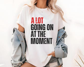 May include: White t-shirt with the text "A LOT GOING ON AT THE MOMENT" in red and black block letters. The shirt is worn with a light blue denim jacket.