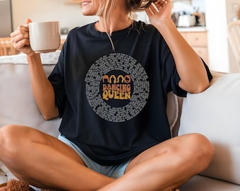 May include: Black t-shirt with a circular design featuring the words "Dancing Queen" in orange and yellow, surrounded by song lyrics. The shirt is worn by a person holding a white mug.