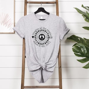 May include: A light gray t-shirt with a black graphic design. The design features a peace symbol surrounded by guitars, hearts, and headphones, with the text "Imagine all the people livin' life in peace."