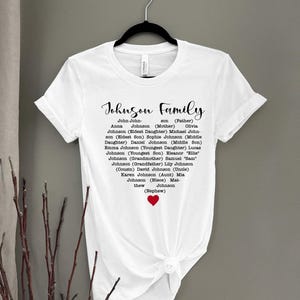 May include: White t-shirt with the words "Johnson Family" in a script font. The names of family members are listed below the title, with a small red heart at the bottom. The shirt is hanging on a black hanger.