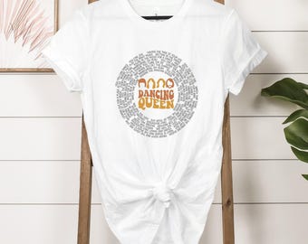 May include: White t-shirt featuring a circular design with the words "Dancing Queen" in orange and brown. The design is surrounded by smaller text. The shirt is tied at the waist.
