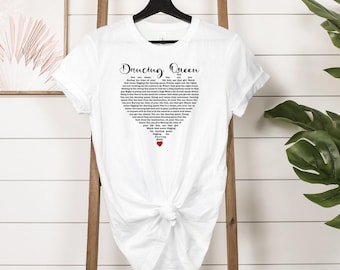 May include: White t-shirt featuring the words "Dancing Queen" in a script font. The lyrics to the song are arranged in a heart shape. A small red heart is at the bottom of the heart-shaped lyrics.