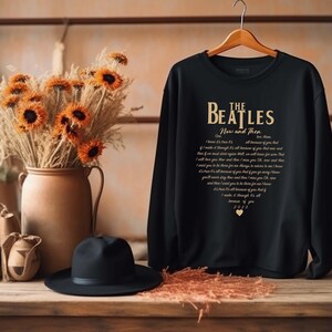 May include: Black sweatshirt with the text "THE BEATLES Now and Then" in gold lettering. The lyrics of the song are printed below the band's name. A black hat and a vase of dried flowers are in the foreground.