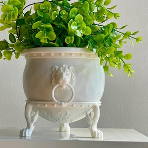 Puede incluir: Maceta de cerámica blanca con detalles de cabeza de león y cuatro patas. La maceta está llena de plantas verdes de hoja.