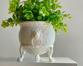 May include: White ceramic planter with lion head details and four legs. The planter is filled with green leafy plants.