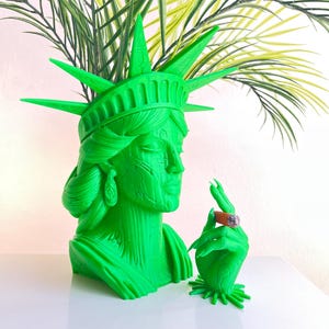 Könnte beinhalten: Ein leuchtend grüner Freiheitsstatue-Blumentopf mit einer Hand, die eine brennende Zigarre hält. Der Topf ist mit künstlichen grünen und gelben Pflanzen gefüllt. Die Statue hat ein detailliertes Gesicht und eine Krone.