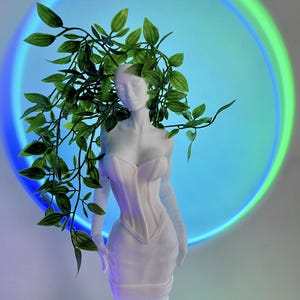 May include: White figurine of a person wearing a white corset and a crown of green leaves. The figurine is standing in front of a blue and green circle of light.