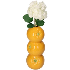 Puede incluir: Un jarrón de cerámica con forma de naranjas apiladas, pintado en un tono naranja brillante. El jarrón contiene un racimo de flores blancas con hojas verdes. El diseño es caprichoso y decorativo, adecuado para la decoración del hogar.