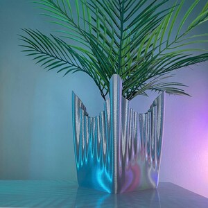 May include: A silver iridescent planter with a unique, wavy design. The planter is filled with a green leafy plant.