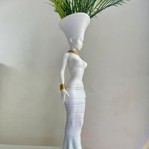 May include: A white decorative vase shaped like a woman, featuring gold details and a tall headdress. The vase contains green and yellow artificial plants. The woman's white dress has a ribbed texture.