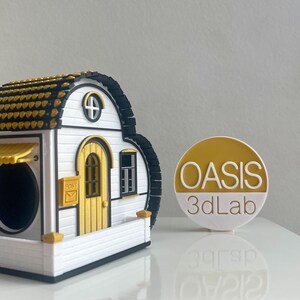 May include: A white and black 3D printed model of a small house with a yellow door and a gold roof. The house has a small window and a mailbox. The model is sitting on a white surface. The text "OASIS 3dLab" is visible on a yellow circle in the background.