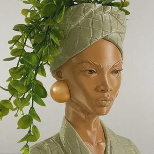 May include: A decorative bust of a woman with a light brown complexion, wearing a quilted sage green head wrap and jacket. The bust features large gold-colored earrings and is adorned with cascading green foliage, creating a unique and artistic display.