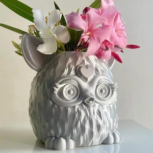 Può includere: Un vaso peloso argentato a forma di animale dei cartoni animati con un naso a forma di cuore e occhi grandi. Il vaso è pieno di fiori rosa e bianchi.