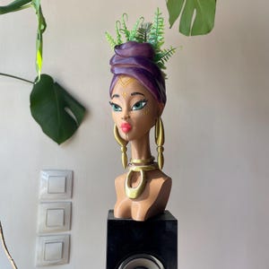May include: A decorative bust of a woman with a purple head wrap and green accents. The bust has gold earrings and a gold necklace. The woman's face is painted with green eyes and red lips. The bust is displayed on a black stand.