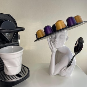 May include: A white ceramic sculpture of a person with a black hat holding coffee pods. The sculpture is holding a black spoon. A white coffee cup and a black espresso machine are in the background.