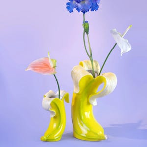 Pode incluir: Dois vasos de cerâmica em forma de banana, um menor que o outro, com exterior amarelo brilhante e interior creme. Cada vaso contém uma única flor: um antúrio rosa, uma flor azul e uma flor branca. O fundo é de cor lavanda lisa.