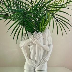 Peut inclure: Un vase en céramique blanc représentant des figures enlacées, contenant des plantes vertes à feuilles. Le vase a une finition brillante et une forme humaine détaillée. Les plantes vertes contrastent avec le vase blanc.