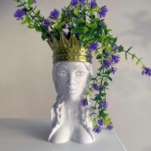 May include: A white ceramic vase in the shape of a woman's head, adorned with a gold crown and braids, filled with purple and green artificial flowers. The vase sits on a white surface.