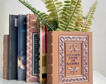 Könnte beinhalten: Ein brauner, buchförmiger Pflanzkasten mit goldenem und rosafarbenem Design. Der Pflanzkasten trägt die Aufschrift "Oxford Book of Nature" auf der Vorderseite. Eine grüne Farnpflanze wächst im Pflanzkasten.