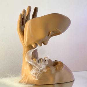 May include: A tan incense holder in the shape of a human face and hand. The face is in profile, with smoke rising from the mouth. The hand supports the face, creating a unique artistic design.