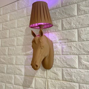 Op de afbeelding: Een bruin gekleurde wandlamp met een paardenkop ontwerp. De lamp heeft een bruine lampenkap en een paars licht. De lamp is gemonteerd op een witte bakstenen muur. Een wit snoer hangt van de lamp af.